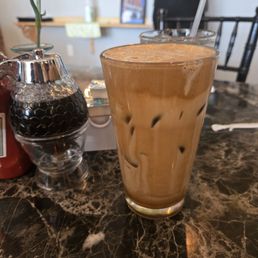 Hazelnut Frappe Iced Coffee - A tall glass of iced coffee with milk and ice cubes is on a marble table in a cafe setting, with a straw in the drink.