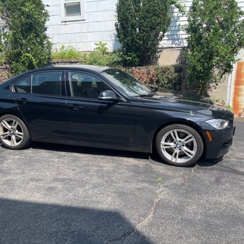 Baltimore Bimmer - A black BMW sedan is parked on a paved surface in an outdoor setting. The vehicle has alloy wheels and tinted windows. Behind the car, there are bushes and trees, along with a white building featuring a window and a rusty metal door. The scene is illuminated by sunlight, casting shadows on the ground. - Auto Repair Near Me - Rosedale, Maryland