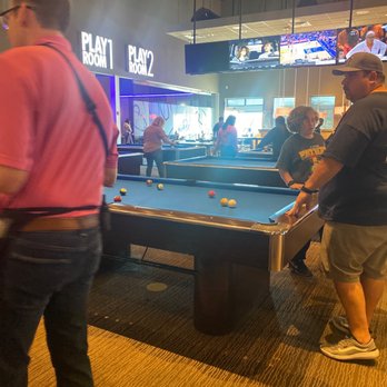 Main Event Gilbert - Several individuals are gathered around a pool table in an arcade or entertainment venue, with screens and game signs visible in the background. - Bowling Near Me - Gilbert, Arizona