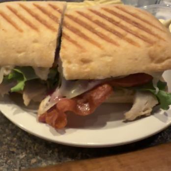 Paradise Pub - Turkey panini - A sandwich featuring grilled bread with visible grill marks, lettuce, tomato, melted cheese, and fried chicken is placed on a white plate. The background shows a glass of water, a small dish with butter, and a dark countertop, indicating a casual dining setting. - Burgers Near Me - Monaca, Pennsylvania