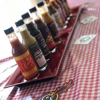 Heatonist - My Hot Ones Challenge with the season line up - The image shows a red tray holding several small bottles of hot sauce with black caps, arranged in a line on a red and white checkered tablecloth. The bottles have various labels, indicating different flavors or brands. There are a few cards and a sticker with a cartoon bomb character on the table. The background is blurred, suggesting an indoor setting with natural light. - Herbs & Spices Near Me - Brooklyn, New York