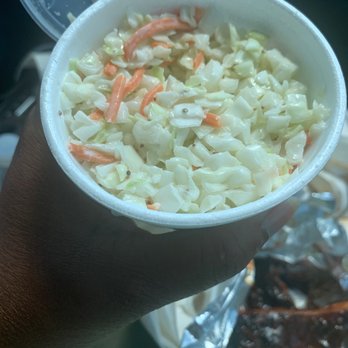 Ray Ray's Hog Pit - Cole Slaw - A person with dark skin is holding a white disposable cup filled with coleslaw, which includes shredded cabbage and carrots. The background shows part of a plastic container with more food and some foil-wrapped items, suggesting a meal or takeout setting. - Barbeque Near Me - Westerville, Ohio