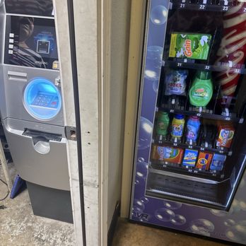 Sunshine Laundry - The image shows a vending machine on the right side, stocked with snacks and drinks, including Gain detergent and other products. To the left, there is a self-service laundry kiosk with a digital screen and keypad, featuring a blue-lit circular area. The setting appears to be inside a laundromat, with a concrete floor and a wall separating the two machines. - Laundromat Near Me - Carmichael, California