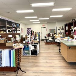 Frugal Fabrics - Photo of the interior of Frugal Fabrics showing fabric and other supplies, as well as one of the proprietors at the cutting table.