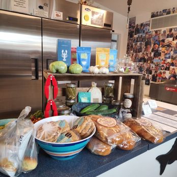 Bennett Shellfish - Counter at a seafood shop featuring baked bread, packaged seafood, fresh vegetables like cucumbers, and jars of preserved items. In the background, a stainless steel refrigerator and a collage of photos on the wall are visible. The display includes various packaged products, with some handwritten labels and a bowl of baked goods. The setting suggests a local seafood and produce shop with a focus on fresh and packaged items. - Seafood Near Me - Montauk, New York
