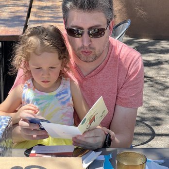 Village Pizza - Great local beer and awesome pizza to go - A man with gray hair, sunglasses, and a pink shirt is seated outdoors with a young girl, both focused on a booklet they are holding. The table in front of them has various papers, a gold-colored glass, and a pizza box. Shadows from nearby trees are visible on the ground and table, indicating a sunny day. - Pizza Near Me - Corrales, New Mexico