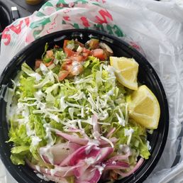 El Pastor Burrito Bowl - A takeout salad in a black container includes lettuce, diced tomatoes, chopped onions, lemon wedges, and dressing, with two small containers of sauce in the background.