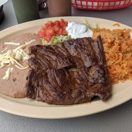 Carne Asada with rice and beans. - A food plate from a Mexican restaurant includes grilled carne asada with a dark, glazed surface, accompanied by rice, refried beans topped with shredded cheese, and garnished with chopped tomatoes, shredded lettuce, sour cream, and salsa. The dish is served on a beige oval plate placed on a gray table, with condiments and utensils visible in the background.