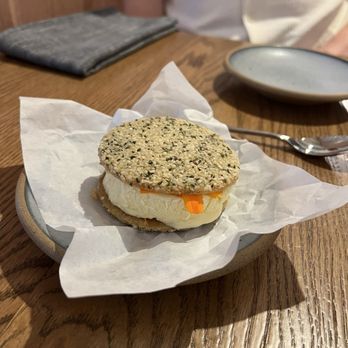 Wildweed - Pawpaw semifreddo sandwich - A sandwich with a seed-covered bun, cheese, and vegetables is served on a plate with parchment paper at a restaurant table. - New American Near Me - Cincinnati, Ohio