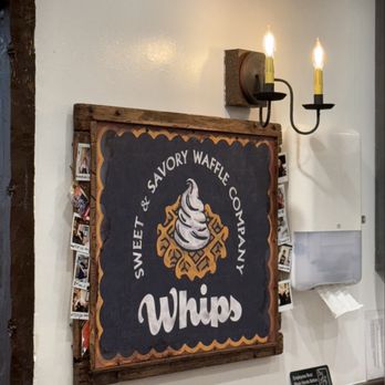 Whips Waffle - A chalkboard sign for Whips, a waffle shop, is displayed on a wall, with a light fixture featuring two candle-shaped bulbs above. - Waffles Near Me - St. Augustine, Florida