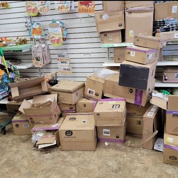 Dollar Tree - Pictures worth a thousand words - The image shows a store interior with a disorganized arrangement of cardboard boxes, some open and others closed, placed on the floor. Shelves in the background hold various products, including candles, decorative items, and small containers. A shopping cart filled with miscellaneous items is positioned on the left side of the image. The floor appears to be made of a textured material, and the overall scene suggests a retail environment with cluttered storage or display areas. - Discount Store Near Me - Pompano Beach, Florida