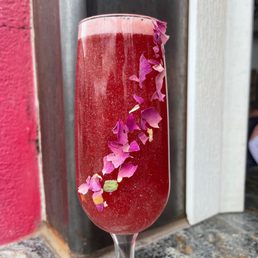 Glitter - A flute glass contains a red drink with purple flower petals floating on top, placed on a textured surface near a wooden wall.