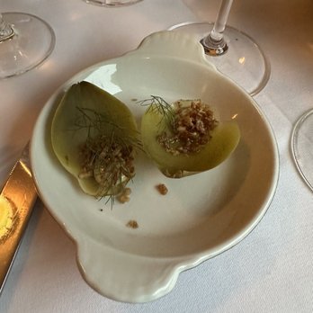 Primo - Amuse bouche - Two halved green grapes with garnishes and toppings are placed on a white ceramic dish, set on a table with wine glasses and a white tablecloth. - Mediterranean Near Me - Rockland, Maine