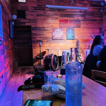 Uncommon Ground - a table with a glass of water and a cell phone on it - A stage setup with musical instruments including a drum, microphone, and stool is positioned against a wooden-paneled wall with signs reading 'Live Music Nightly' and 'Uncommon Ground' at Uncommon Ground in Chicago. The scene includes a table in the foreground with a water bottle, glass, plates, and a camera, indicating a casual dining or event setting. The lighting has a purple hue, and a person is partially visible on the right side of the image. - Breakfast & Brunch Near Me - Chicago, Illinois