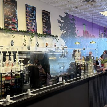 Wild Pine - The image shows the interior of Wild Pine Coffee, featuring a counter where customers place their orders. A barista is preparing drinks behind the counter, which is equipped with coffee-making supplies. Several customers are standing in line, some wearing masks, and a mural depicting a landscape with trees and mountains decorates the wall behind the counter. Overhead lighting and a small potted plant add to the ambiance. - Coffee & Tea Near Me - Peoria, Arizona