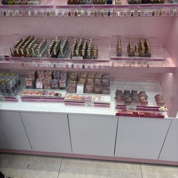 Tokimo - The image features a retail display of lip cosmetics, including lipsticks and lip glosses, arranged on pink shelves within a makeup store. The products are in metallic and transparent packaging, organized in rows and sections. The display is part of a larger makeup section, with additional cosmetic products visible in the background. The store has a modern, clean aesthetic with a pink color theme and white cabinetry at the bottom of the display. - Cosmetics & Beauty Supply Near Me - Flushing, New York