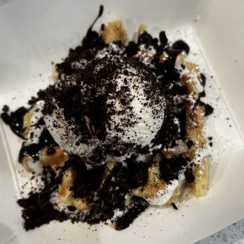 Wondaffle - Cravin Cookies Leige waffle combo - A dessert served in a white paper container, consisting of a waffle topped with a scoop of ice cream, drizzled with chocolate syrup, and sprinkled with cookie crumbs. The waffle appears to be golden brown, and the ice cream is partially covered by the toppings. The background shows a speckled gray surface. - Waffles Near Me - Bethlehem, Pennsylvania