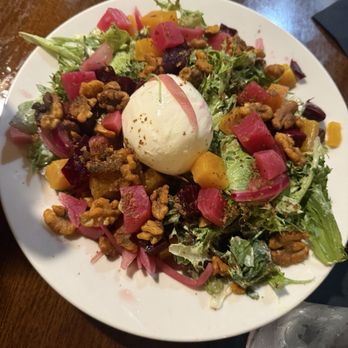 The Mill - Hershey - Beet and Burrata Salad - A salad featuring mixed greens, diced pickled vegetables in pink, yellow, and purple, sprinkled walnuts, and a poached egg in the center, served on a white round plate. The dish is placed on a wooden table with a glass of water nearby. - New American Near Me - Hershey, Pennsylvania