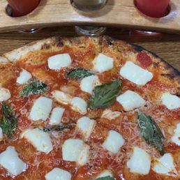 Margherita - A pizza Margherita with basil leaves and chunks of mozzarella cheese is displayed on a round tray. Above the pizza, three small shot glasses contain different colored beverages, placed in a wooden holder. The setting appears to be a restaurant or brewery with a wooden table surface.