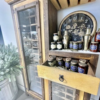 Daily Blooms - Scented candles - A wooden display cabinet with glass doors and a rustic finish is positioned against a wall. On top of the cabinet, there is a vintage-style clock with visible gears and a black face. Nearby, there are several jars and containers, some with labels, arranged on the cabinet's surface. To the left, a large potted plant with green foliage is visible. The setting appears to be indoors, possibly in a store or a home with decorative items. - Home Decor Near Me - Fallbrook, California