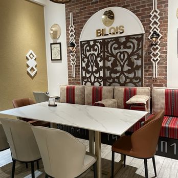 Bilqis Coffee - The interior of Bilqis Coffee includes a white marble table surrounded by different chairs, including beige, brown, and black options. The background features a brick wall with decorative elements, such as a large sign reading 'BILQIS,' ornate metalwork, and wall-mounted lanterns. An air conditioning unit is visible above the sign, and the space is lit with ceiling lights and a hanging lantern. The decor combines modern and traditional design elements. - Coffee & Tea Near Me - Brooklyn, New York