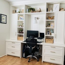 Custom office built in - A custom white office built-in unit with multiple open shelves, closed cabinets, and drawers is shown. The shelves contain decorative items, framed photos, and small plants. A desk with a computer monitor, laptop, and office chair is positioned in front of the unit. The setup is situated against a light-colored wall with a wooden floor, creating an organized workspace.