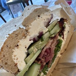 Wilson Ave (the best bagel here) - A bagel sandwich filled with cream cheese, cucumber slices, and meat, placed on parchment paper inside a cafe or bakery.