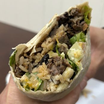 Taco Mogo - Burrito w/bulgogi - A partially eaten burrito filled with rice, ground beef, lettuce, black beans, and other ingredients, held in a person's hand. The burrito's tortilla is visible, and the filling is exposed, showing a mix of textures and colors. The background is blurred, focusing on the food item. - Korean Near Me - Eugene, Oregon