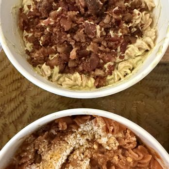 Pastissimo - The image features two bowls of pasta placed on a wooden surface. The top bowl contains pasta with a creamy sauce and is topped with cooked bacon bits. The bottom bowl has pasta with a tomato-based sauce, garnished with breadcrumbs. Both bowls are filled with different pasta dishes, showcasing distinct toppings and sauces. - Italian Near Me - Austin, Texas