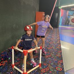 Classic skating! - Two children roller skate on a colorful patterned floor inside an entertainment center, one wearing a Spider-Man helmet and the other with a helmet and a purple shirt.