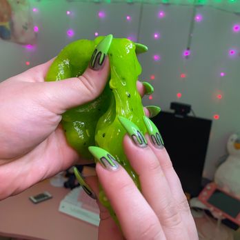 Tip Top Nails - acrylic nails, nail art, green, slime, color. - A person’s hand with long, decorated nails painted in green and black holds a green, slime-covered object. The background features colorful string lights, a plush penguin, and other plush toys, with a pink desk and a small TV or monitor. The scene appears to be indoors with decorative lighting. - Nail Salons Near Me - Sandy, Utah