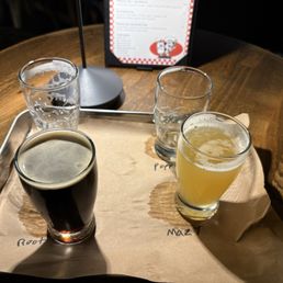 Flight of beers - Four glasses containing different beverages are arranged on a tray lined with paper on a wooden table. The drinks include water, a dark beer, and two light-colored beverages, possibly juice or craft beer. A menu is visible in the background, standing upright, with a black frame and a red and white design. The setting appears to be a restaurant or bar environment.