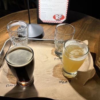 Farm.One - Flight of beers - Four glasses containing different beverages are arranged on a tray lined with paper on a wooden table. The drinks include water, a dark beer, and two light-colored beverages, possibly juice or craft beer. A menu is visible in the background, standing upright, with a black frame and a red and white design. The setting appears to be a restaurant or bar environment. - Farms Near Me - Brooklyn, New York