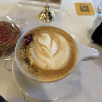 Criterion coffee - pistachio vanilla latte - A latte with foam art, featuring a heart-shaped design, is served in a white ceramic cup on a matching saucer. The cup is on a marble table, with a small packet of cookies partially visible on the side. In the background, there are menu signs and a QR code, indicating a coffee shop setting. - Coffee & Tea Near Me - Pasadena, California