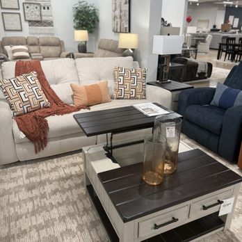 Ashley Store - Furniture store showroom with various sofas, including a beige sofa with patterned pillows, a dark blue armchair, and a beige sectional in the background. There are side tables with lamps, decorative vases, and wall art. The space is well-lit with ceiling lights and has a neutral-colored carpeted floor. - Furniture Stores Near Me - Hunt Valley, Maryland