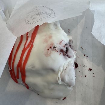 The Crafted Scone - A partially eaten scone with white icing and red drizzle is inside a bakery paper wrapper. The scone has a rough, crumbly texture with some crumbs and red specks visible around it. The wrapper has a logo or text printed on it, indicating it is from a bakery or café. The background is plain and neutral. - Bakeries Near Me - Temecula, California