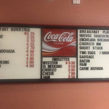 Casa De Benavidez - The image shows a menu board at Casa De Benavidez in Albuquerque, NM, with sections for breakfast burritos, plates, and omelettes. The menu includes items like bacon, sausage, ham, cheese, chicharrone, chorizo, and various breakfast plates such as huevos rancheros and chorizo breakfast. Prices are listed next to each item, with some items featuring multiple price options. The board also displays a Coca-Cola logo in the center. The background wall is painted in a reddish color. - Mexican Near Me - Albuquerque, New Mexico