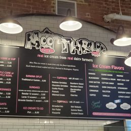 menu - Menu board at Moo Thru ice cream shop listing flavors, toppings, and prices, with a brick wall background and overhead lighting.