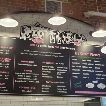 Moo Thru - menu - Menu board at Moo Thru ice cream shop listing flavors, toppings, and prices, with a brick wall background and overhead lighting. - Ice Cream & Frozen Yogurt Near Me - Charlottesville, Virginia