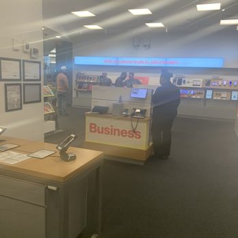 Verizon - Still around like mannequins - The image depicts the interior of a Verizon store, with several customers and staff members visible. A customer stands at a service counter labeled 'Business,' while others are engaged in conversations or browsing. The store features display shelves with electronic devices and accessories, illuminated by bright ceiling lights. The photo is taken through a glass window, with reflections and a slight glare present. - Mobile Phones Near Me - Brooklyn, New York