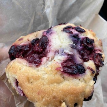 Scratch Kitchen - Blueberry Muffin! - A close-up image of a blueberry muffin with blueberries on top, wrapped in crumpled parchment paper. The muffin has a golden-brown crust with visible blueberries embedded in the top and sides. The interior appears moist and fluffy, with some blueberries slightly bursting. The muffin is presented in a casual setting, emphasizing its baked texture and fresh fruit ingredients. - Breakfast & Brunch Near Me - Olney, Maryland