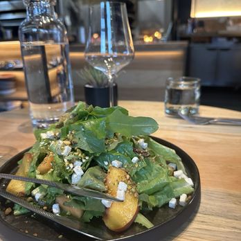 Two Twelve - Salad - A salad featuring mixed greens, peach slices, and crumbled cheese is presented on a black plate placed on a wooden table. The table setting includes a fork, a glass of water, a bottle of water, a wine glass, and a small potted plant in the background. The restaurant interior appears modern with metallic and wooden elements, and ambient lighting is visible. - New American Near Me - Crested Butte, Colorado