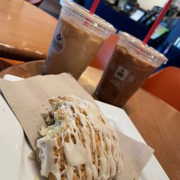Honey Bee - A cinnamon roll with icing on a white plate and a piece of parchment paper, with two iced coffee drinks in the background at a coffee shop.