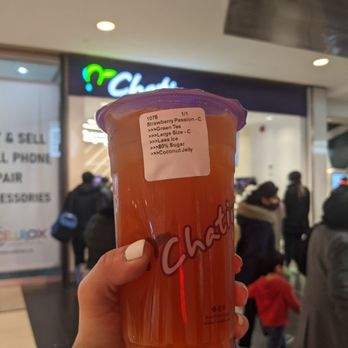 Chatime - Strawberry passion fruit green tea with coconut jelly - A hand with white nail polish holds a large plastic cup of strawberry passion fruit green tea with coconut jelly, labeled with product details. The background shows a shopping mall interior with a Chatime store sign and several shoppers walking around, some wearing winter clothing. The setting indicates a busy retail environment. - Bubble Tea Near Me - Toronto, Ontario