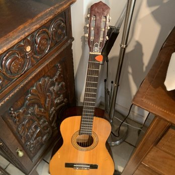 Sonbobs - a guitar in a corner of a room - An acoustic guitar with a natural wood finish is leaning against a wall, with a pick-shaped capo attached to the neck. The guitar is situated next to a carved wooden cabinet and a tall floor lamp, in an indoor setting with tiled flooring. - Coffee & Tea Near Me - Astoria, New York