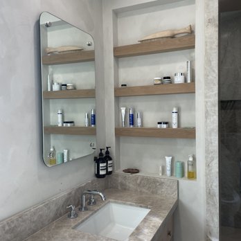PCD Remodeling - The image shows a bathroom vanity with a beige marble countertop and a rectangular white sink. A chrome faucet and handles are installed on the countertop. Above the sink, a wall-mounted mirror with rounded edges reflects part of the room. To the right, built-in recessed shelves with wooden surfaces hold various toiletries, skincare products, and decorative items, including a wooden fish sculpture. The wall behind the shelves and mirror is painted in a light, neutral color, creating a clean and minimalist aesthetic. - General Contractors Near Me - La Jolla, California