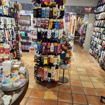 Atown - A gift shop interior features a central rotating display filled with colorful socks, including cartoon characters like SpongeBob and other patterns. The shop has tiled flooring and shelves on both sides stocked with accessories, home decor, and novelty items. Additional displays and racks are visible in the background, with a sign indicating the toilet area. The shop appears well-organized with a variety of locally-made and handmade products. - Accessories Near Me - Austin, Texas