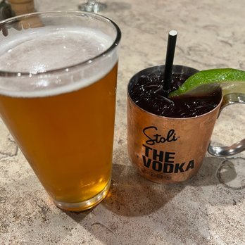 Amaya - Drinks - The image shows a tall, clear glass filled with a golden-colored beer with a foamy head, positioned on a textured, light-colored table. Next to it is a copper-colored mug labeled 'Stoli THE VODKA,' containing a dark cocktail with ice, garnished with a lime wedge and a black straw. The setting appears to be a casual dining or bar environment. - New American Near Me - Santa Fe, New Mexico