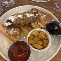 Chicken and Waffles - A breakfast dish with fried chicken placed on top of waffles, dusted with powdered sugar, accompanied by a small bowl of ketchup, a bowl of syrup, and a side of seasoned potato wedges. The plate is on a wooden table, with glasses of water, lime wedges, and a fork visible in the background.