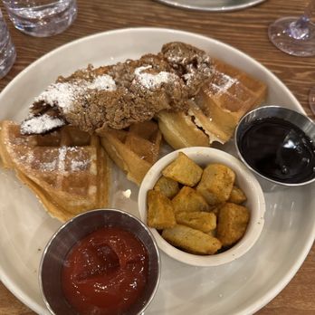 Johnny Pistolas - Chicken and Waffles - A breakfast dish with fried chicken placed on top of waffles, dusted with powdered sugar, accompanied by a small bowl of ketchup, a bowl of syrup, and a side of seasoned potato wedges. The plate is on a wooden table, with glasses of water, lime wedges, and a fork visible in the background. - Mexican Near Me - Washington, DC, District of Columbia